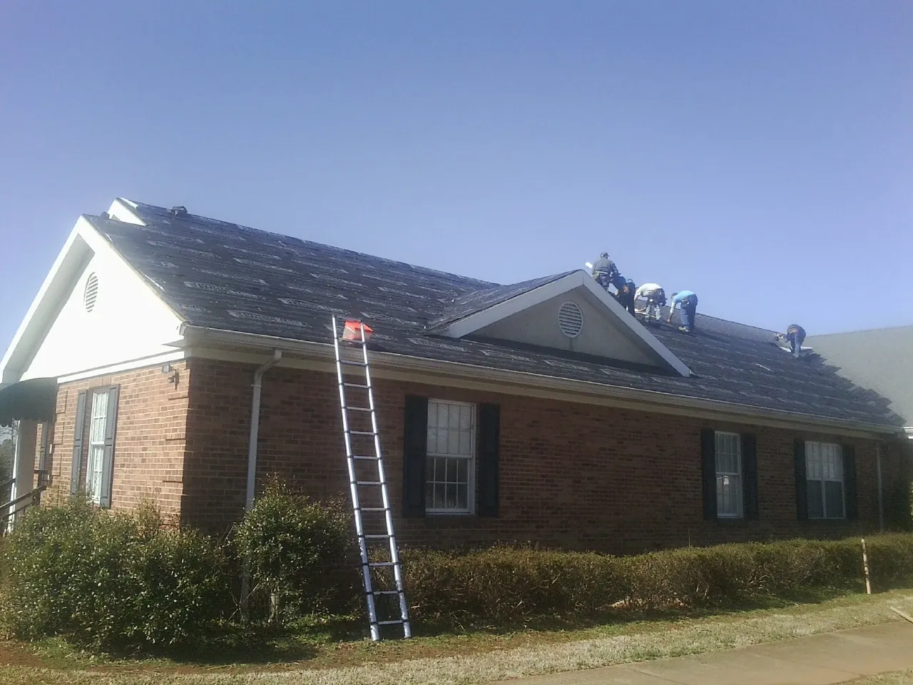 Crew tearing shingles off a brick ranch roof with a ladder set — Metal Roof Repair work by our crew in Castella