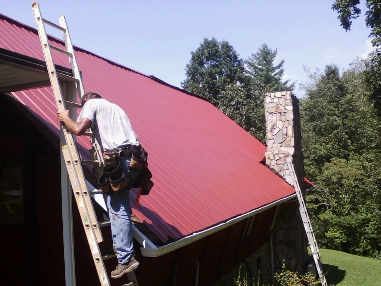 Roofer on a ladder beside a red metal roof and stone chimney — Soffit & Fascia Repair by Roofing Brothers in Castella
